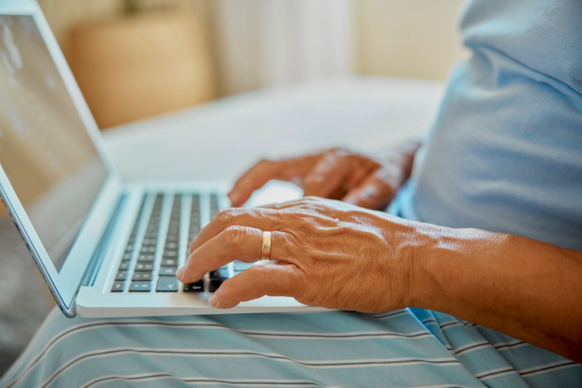 Mani di una persona anziana che digitano su un computer portatile in grembo. Indossano un anello, in casa, probabilmente sul letto.