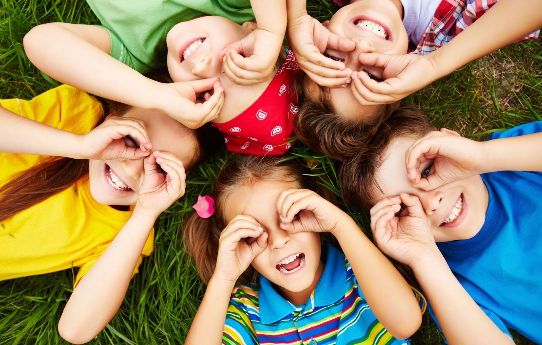 Children lying on grass, forming circles around their eyes with their fingers, smiling.