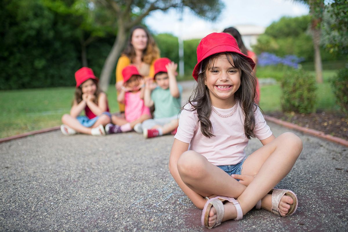Girl with red hat smiles at the camera, sitting on path; other children and adult in background.