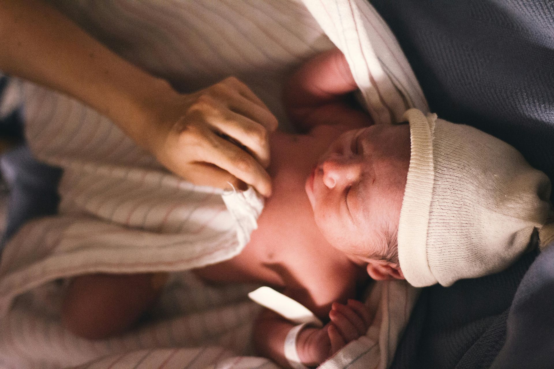 Newborn baby wrapped in a blanket, wearing a hat, with a hand gently adjusting the blanket.