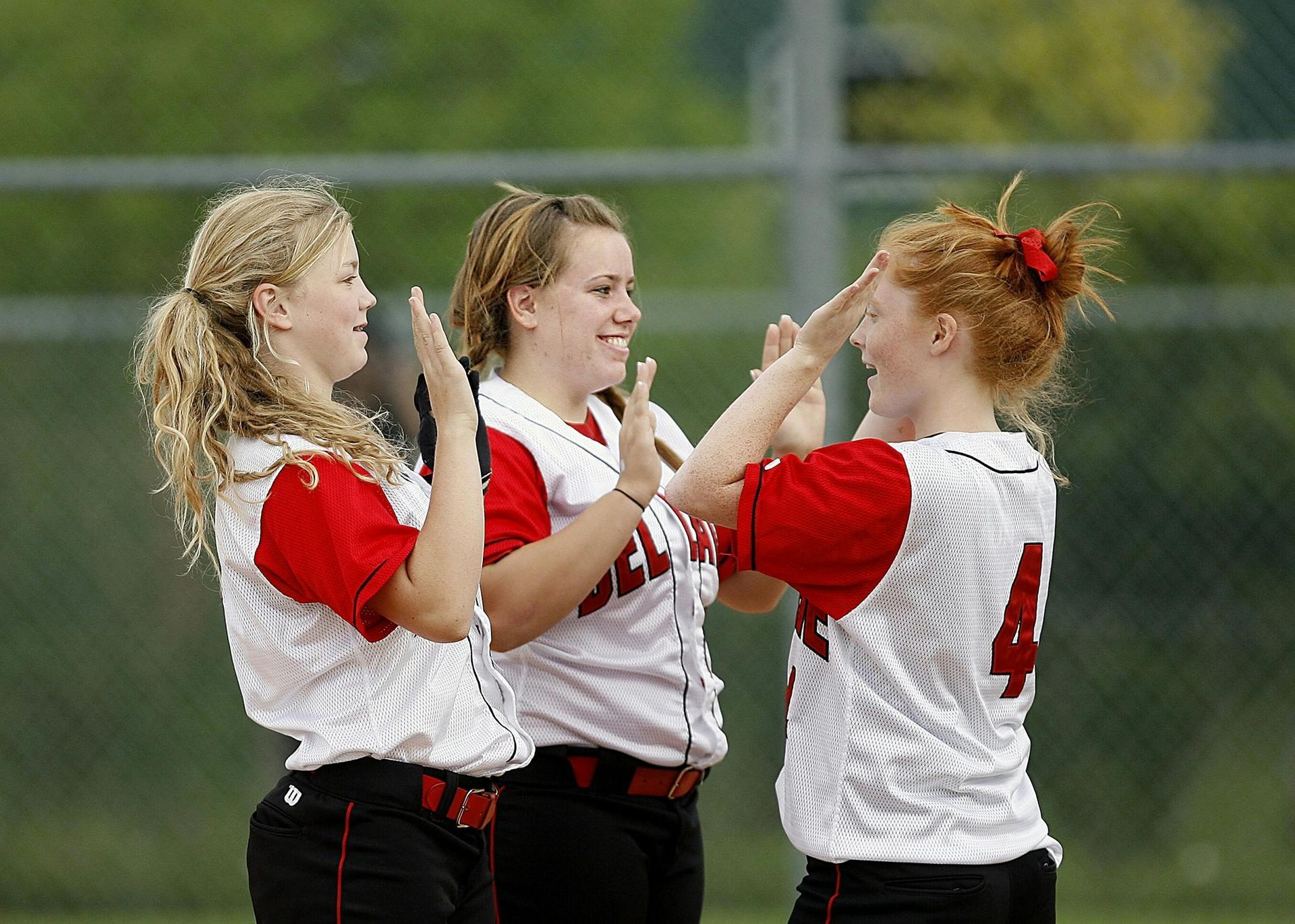Three softball players in red and white uniforms high-fiving on a field.