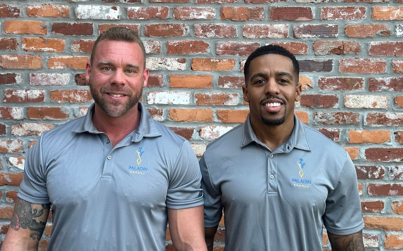 Two men are standing next to each other in front of a brick wall.