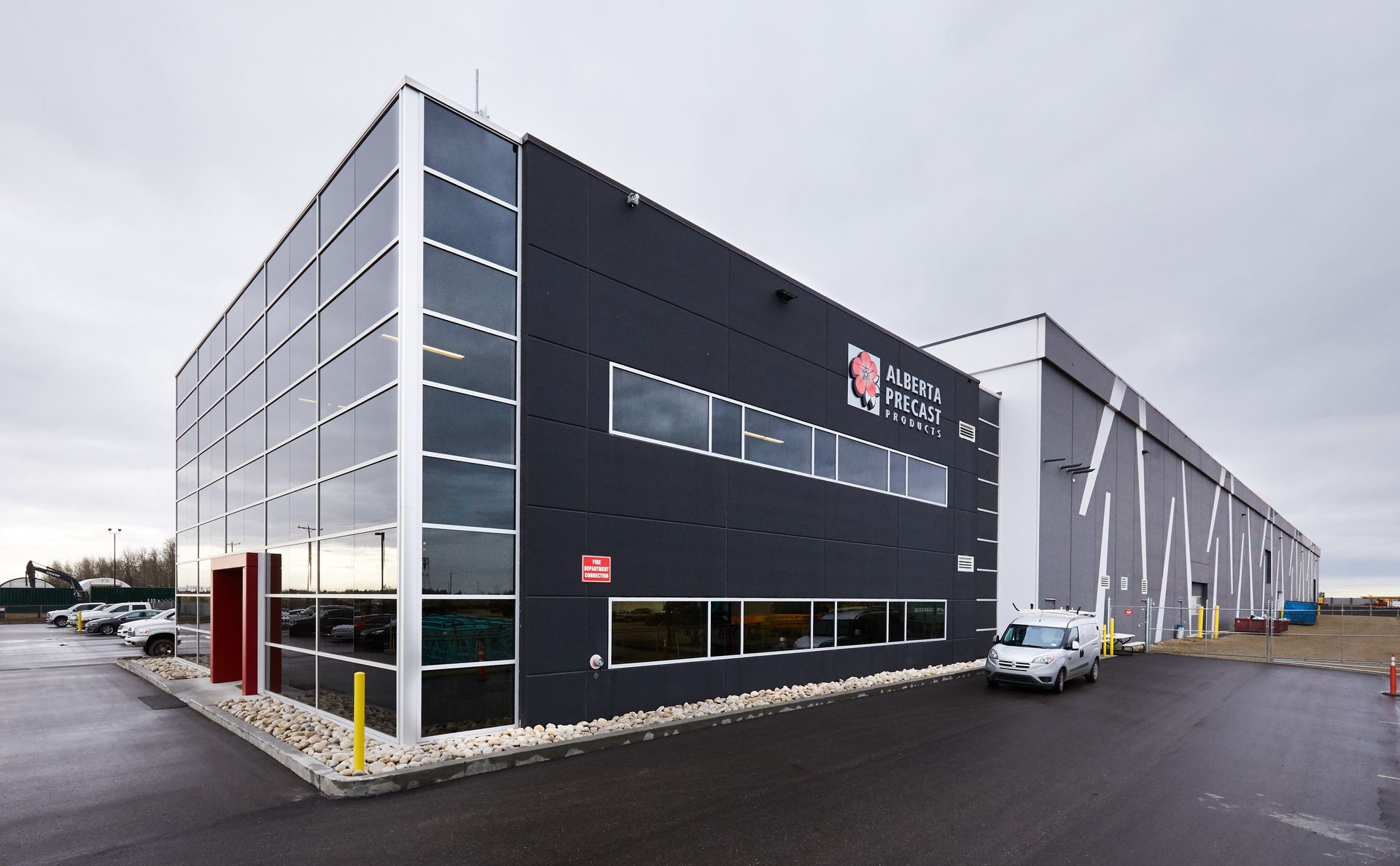 Modern industrial building with black and glass facade, logo, and parking lot. Cloudy sky overhead.