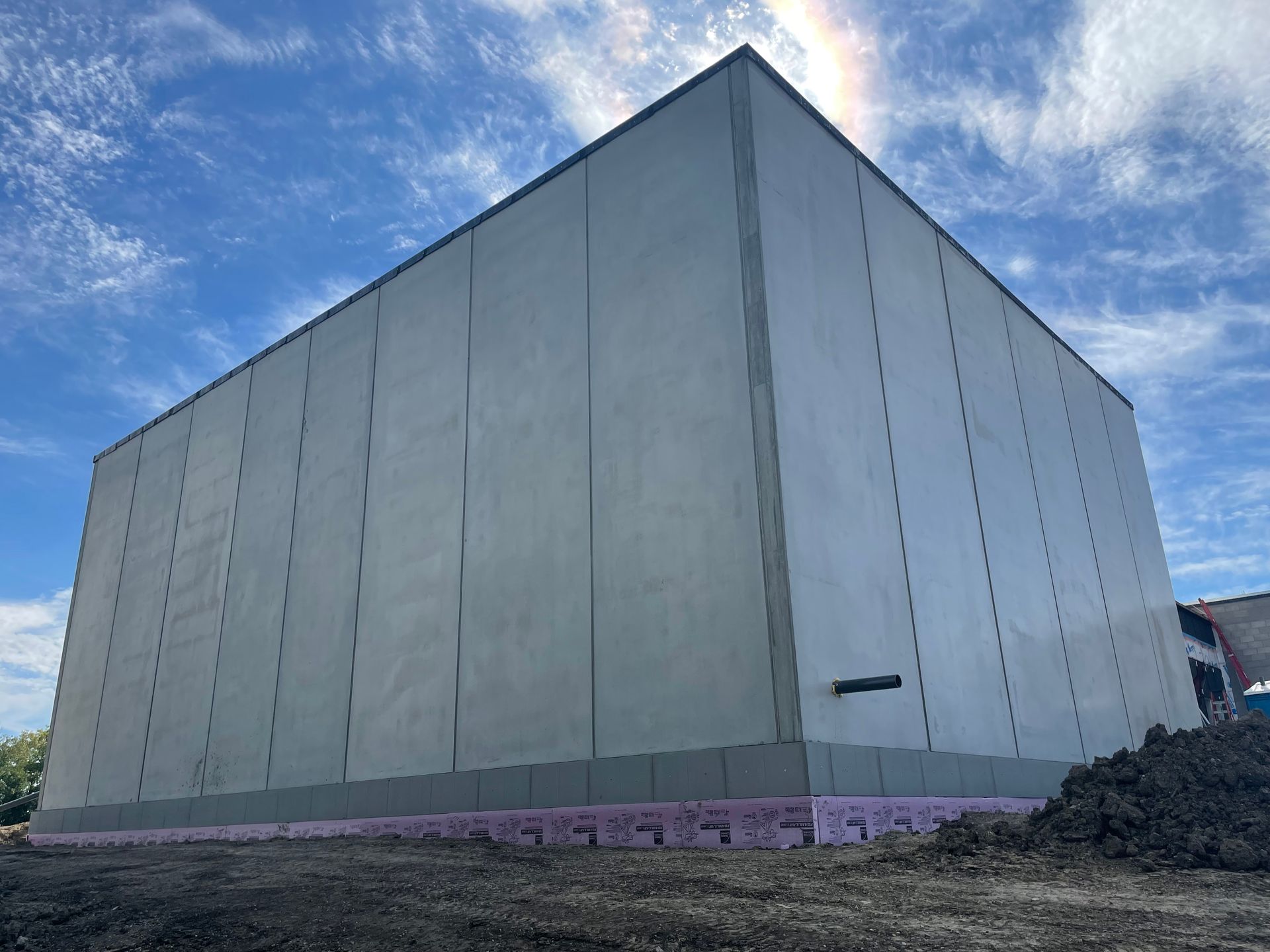 Large, gray industrial building under construction with pink insulation visible at the base against a cloudy blue sky.