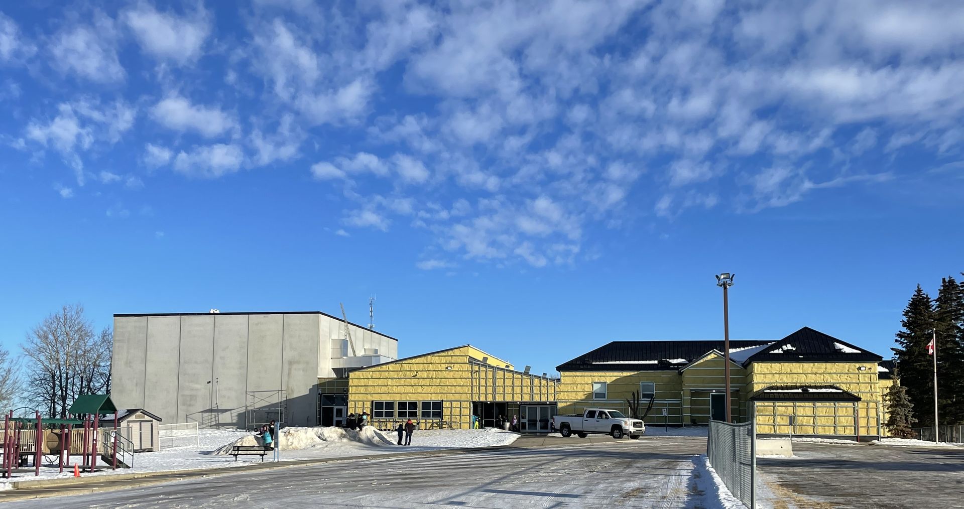 School buildings under a bright blue sky with scattered clouds, snow on the ground and a playground in view.