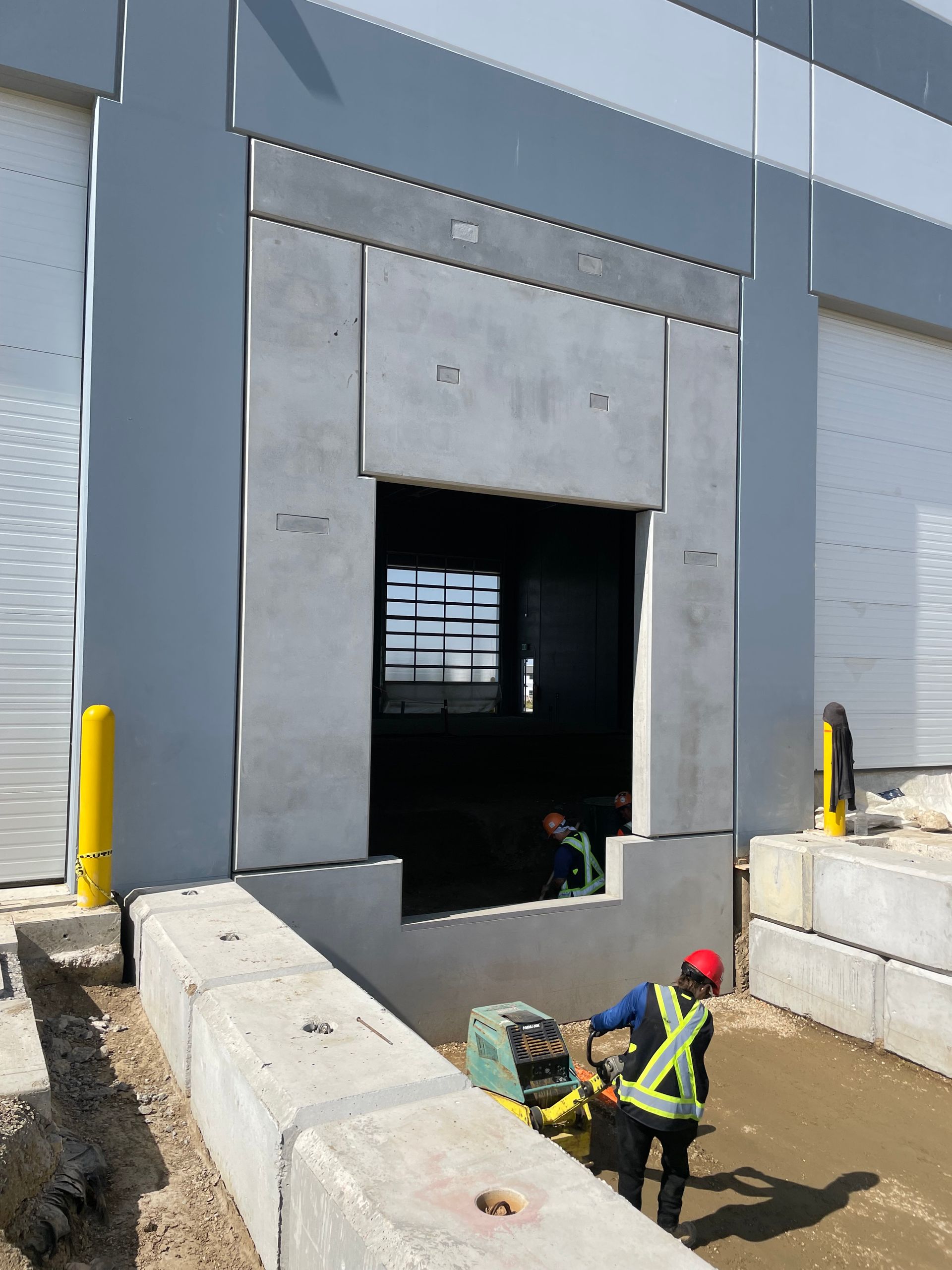 Construction worker compacts ground near a loading dock. Concrete panels frame the opening, with a yellow bollard visible.