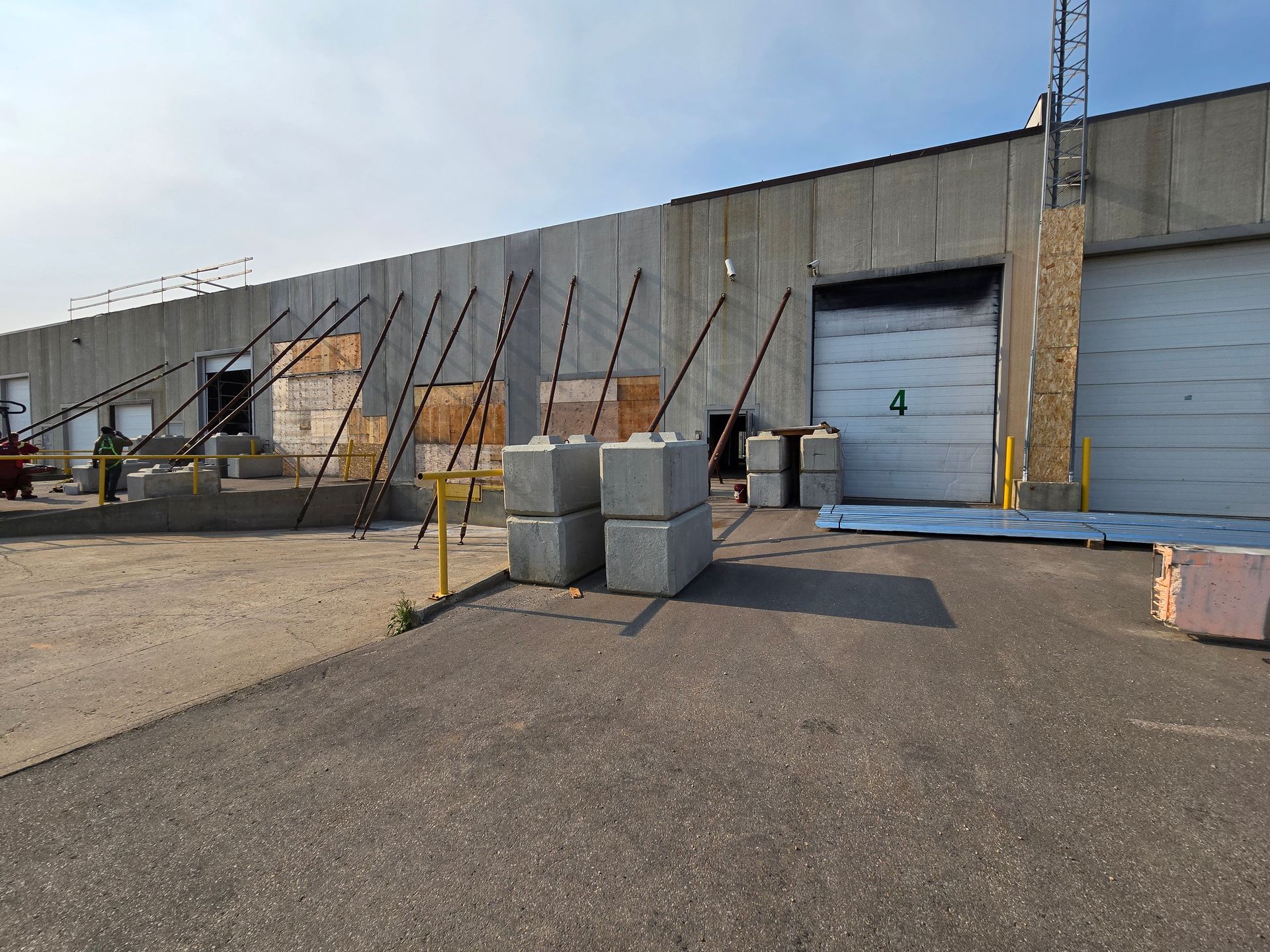 Exterior of a warehouse with support beams bracing the concrete walls near loading docks. Gray concrete blocks are stacked in the foreground.