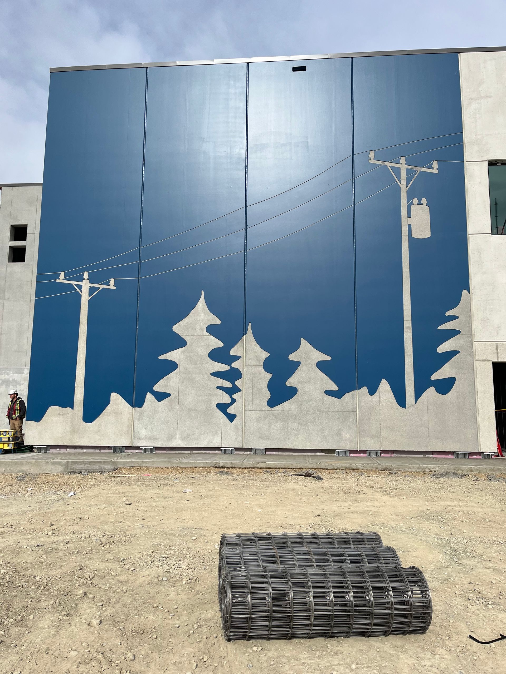 Mural on a building depicting a blue sky, silhouetted trees and utility poles. A pile of black mats lies in front.