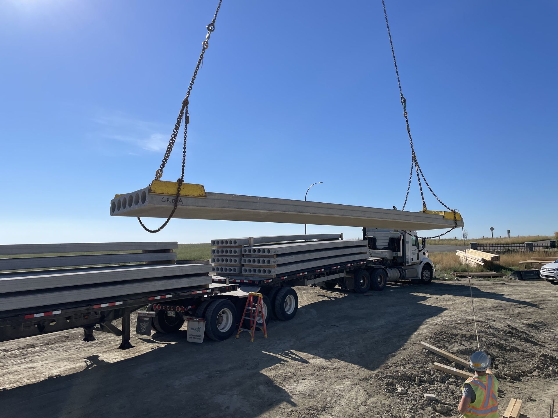 A crane lifting a long concrete slab from a flatbed truck on a construction site. Bright blue sky overhead.