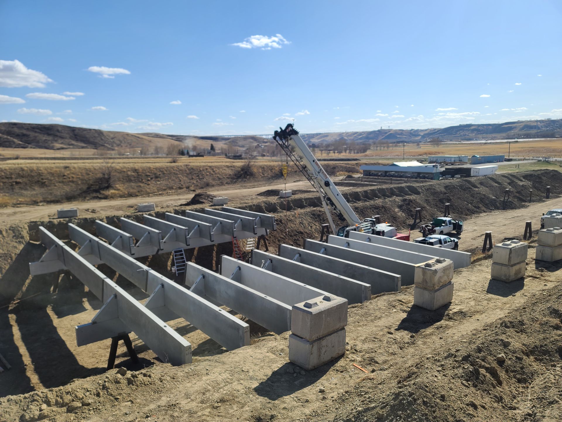 Construction site with a crane placing concrete beams over a ditch. Gray beams and blocks, blue sky, and a brown landscape.