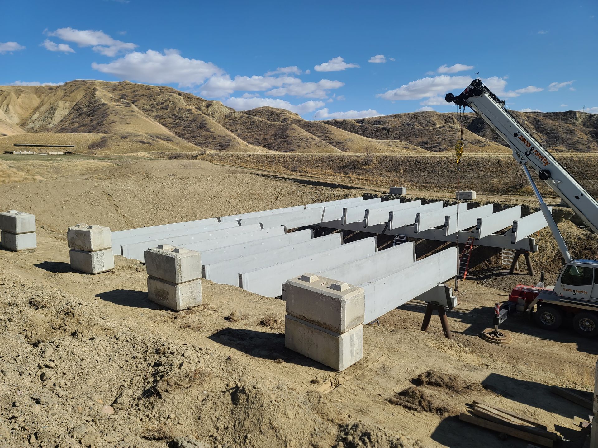 Construction site with a crane assembling concrete beams and blocks on a brown field, with hills in the background.