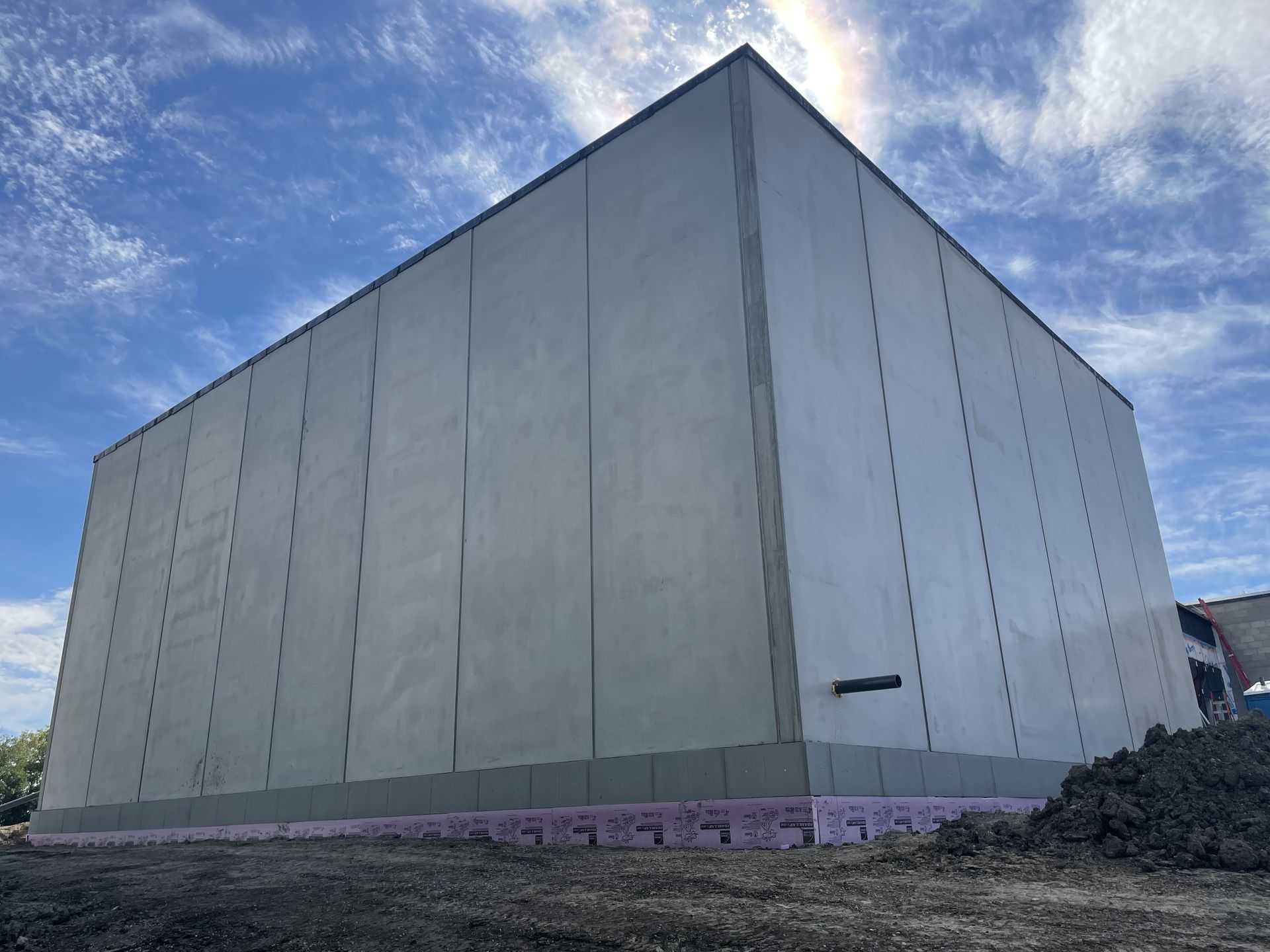 Gray industrial building under construction against a cloudy blue sky. Foundation shows pink insulation.