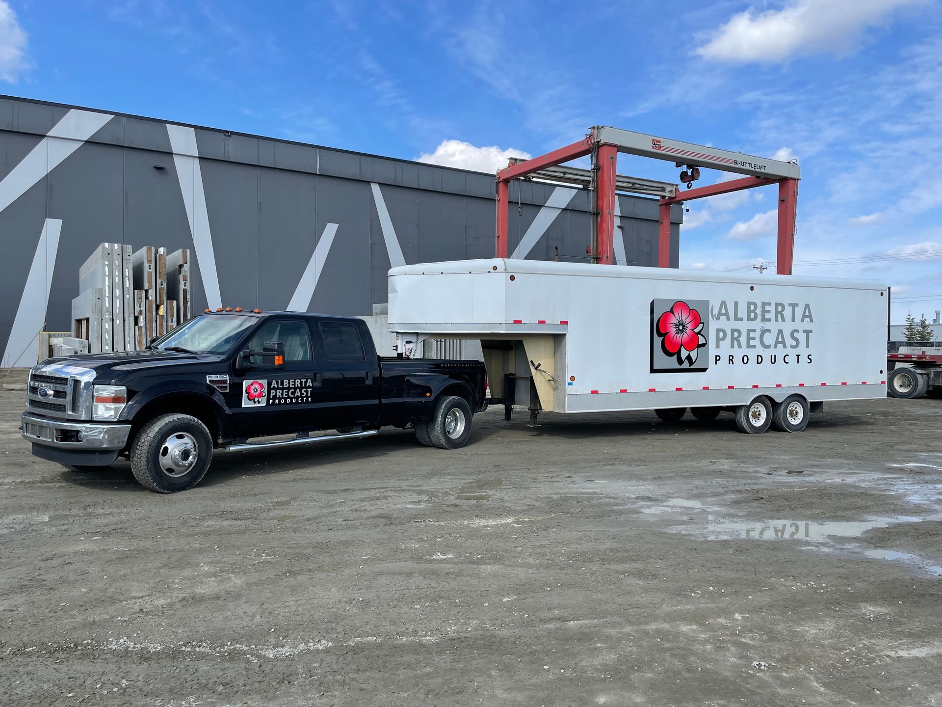 A black pickup truck towing a white trailer with the Alberta Precast logo in front of a building on a gravel lot.