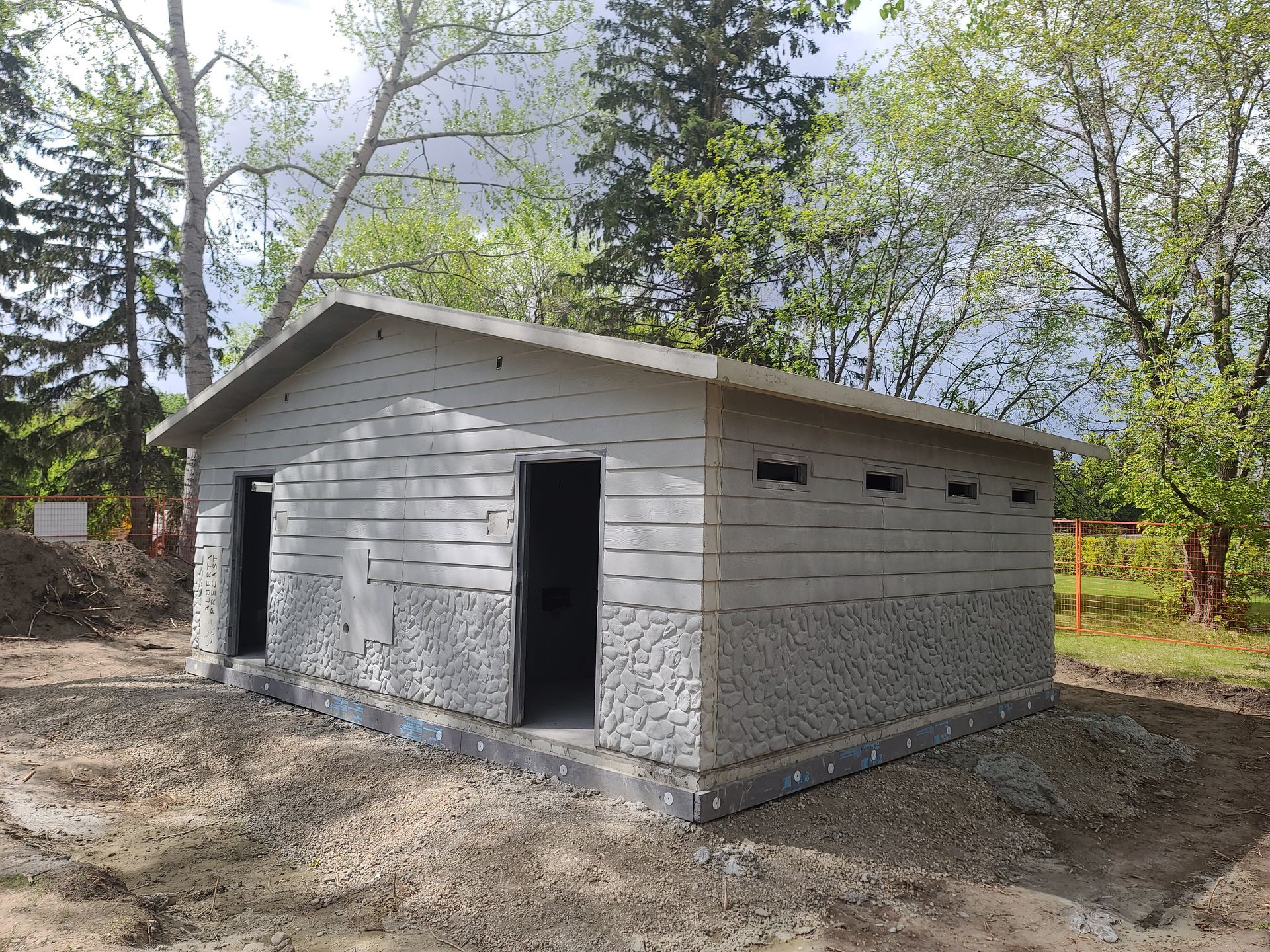 A prefabricated concrete building with two doorways, under construction outdoors. The walls have a textured pattern and the roof is sloped.