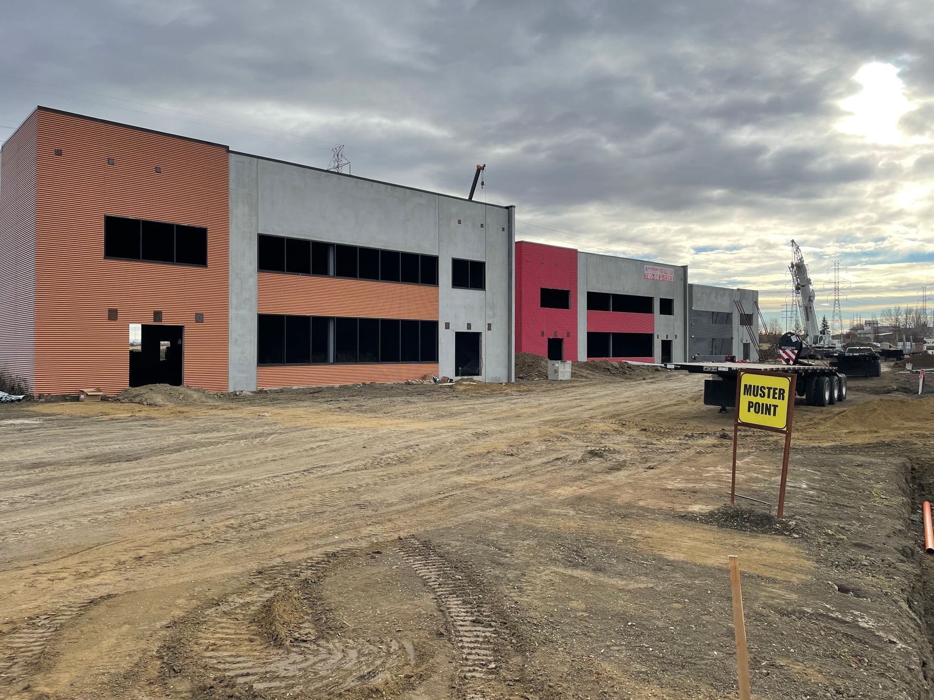 Construction site with a row of industrial buildings, some with colorful murals, under a cloudy sky. A sign on the foreground mentions a project.