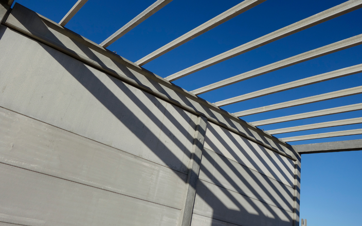 Gray concrete wall with shadow stripes from a white metal pergola against a bright blue sky.