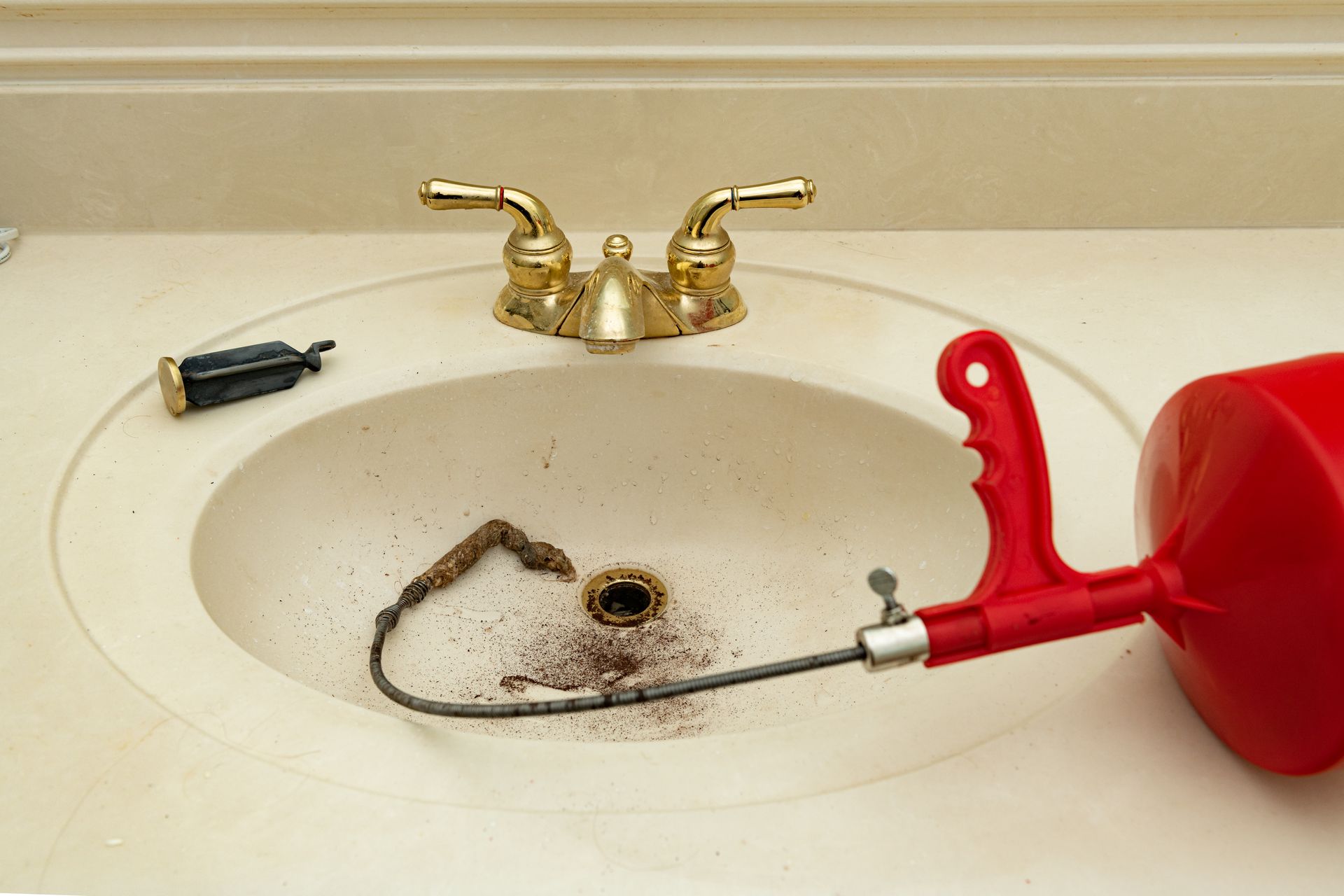 A clogged bathroom sink with a drain snake and a red plunger next to it.
