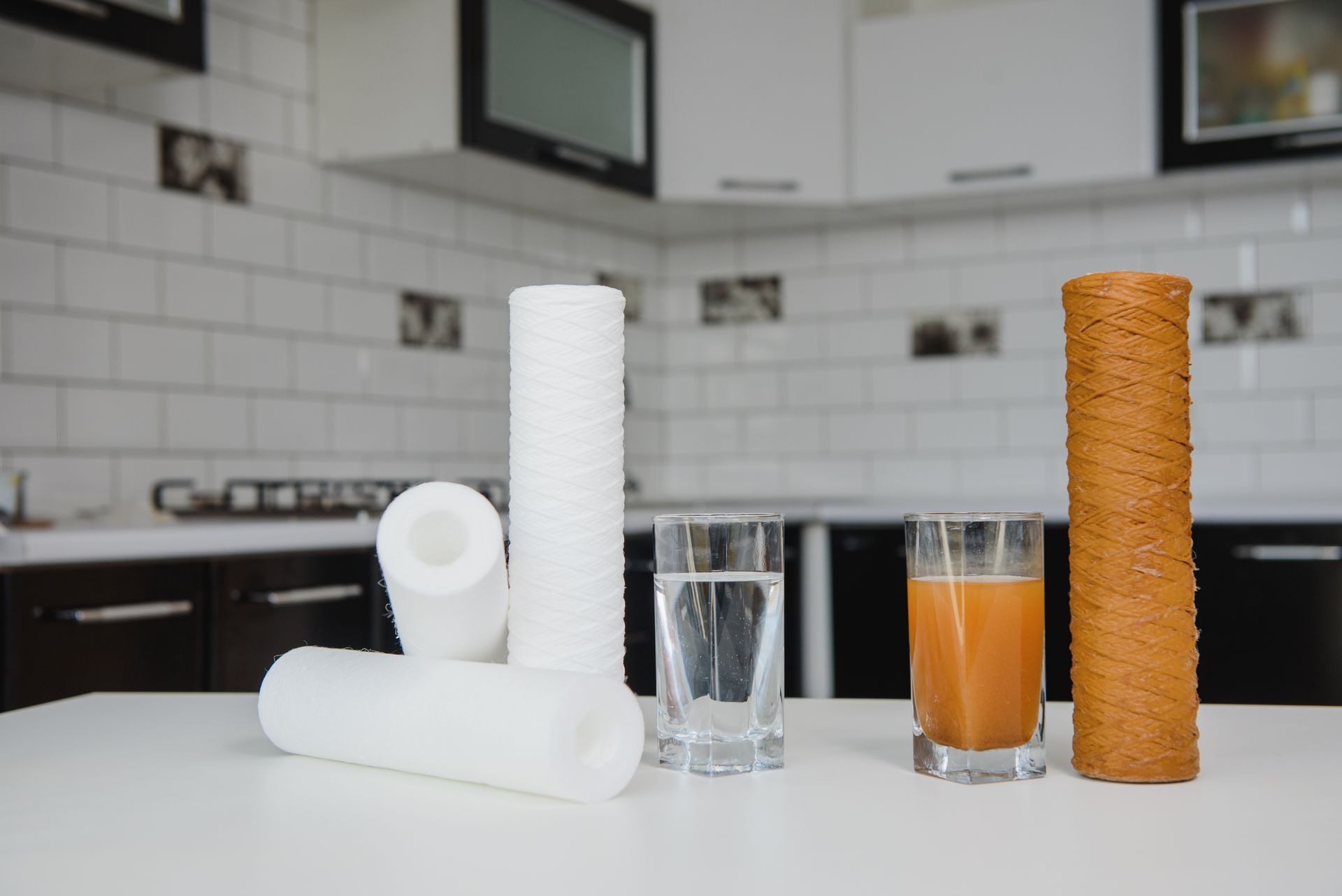 Water filters and two glasses of water, one clear, one brown, on a white table in a kitchen.