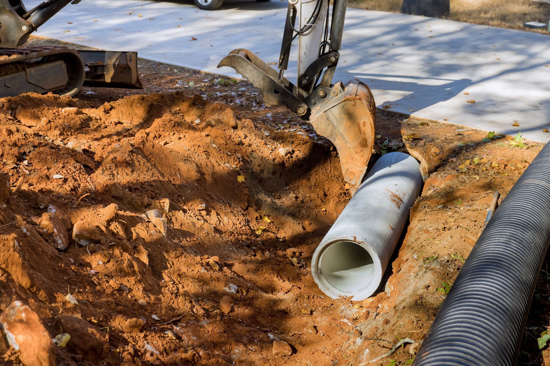 Excavator digging near concrete pipe and corrugated black pipe in dirt, outdoors.