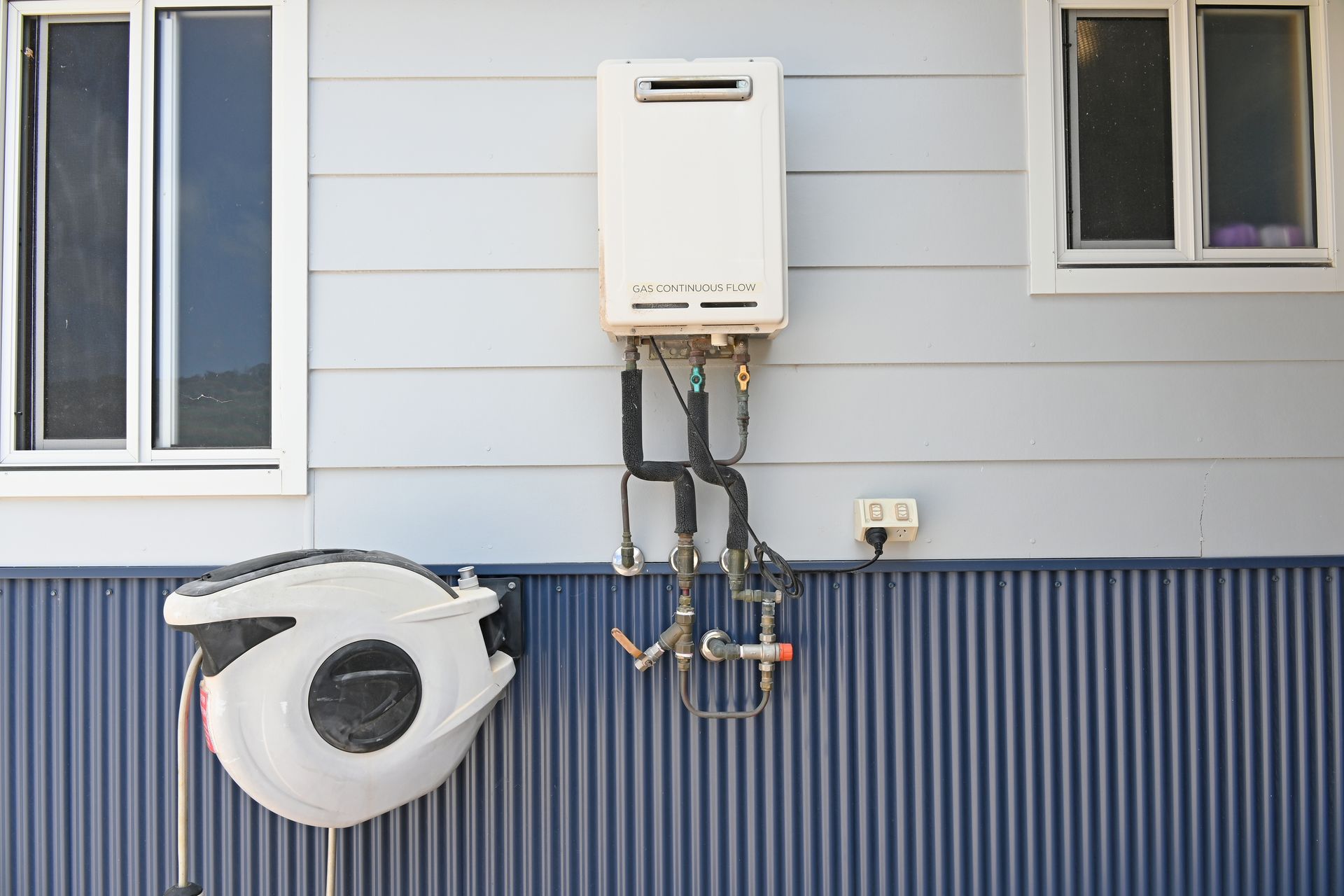 Outdoor water heater mounted on a blue wall between two windows, with hose reel below.