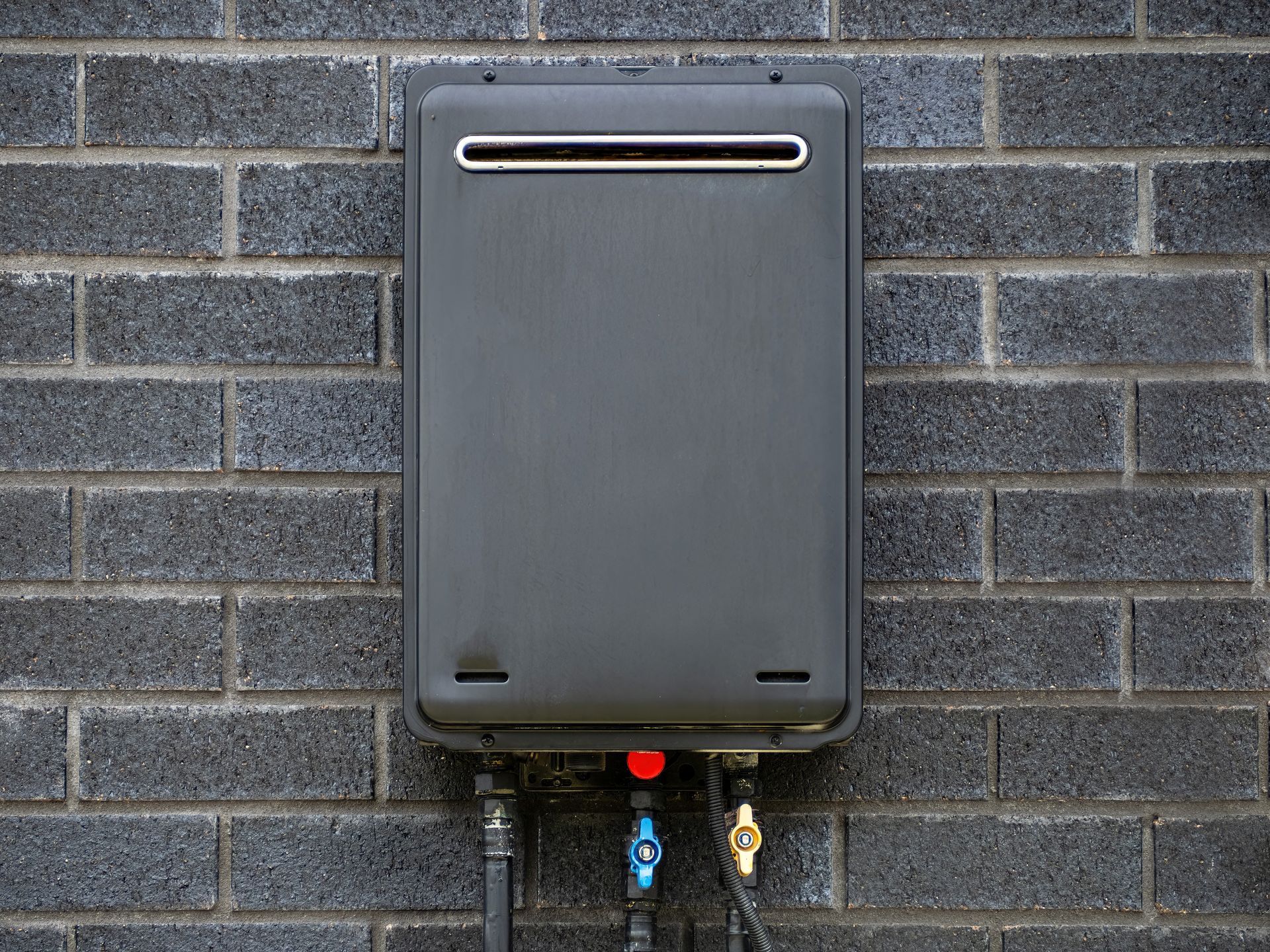 Dark gray tankless water heater mounted on a dark brick wall, with plumbing below.