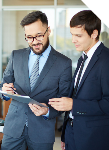 Two men in suits, one pointing at a clipboard, discussing in an office setting.