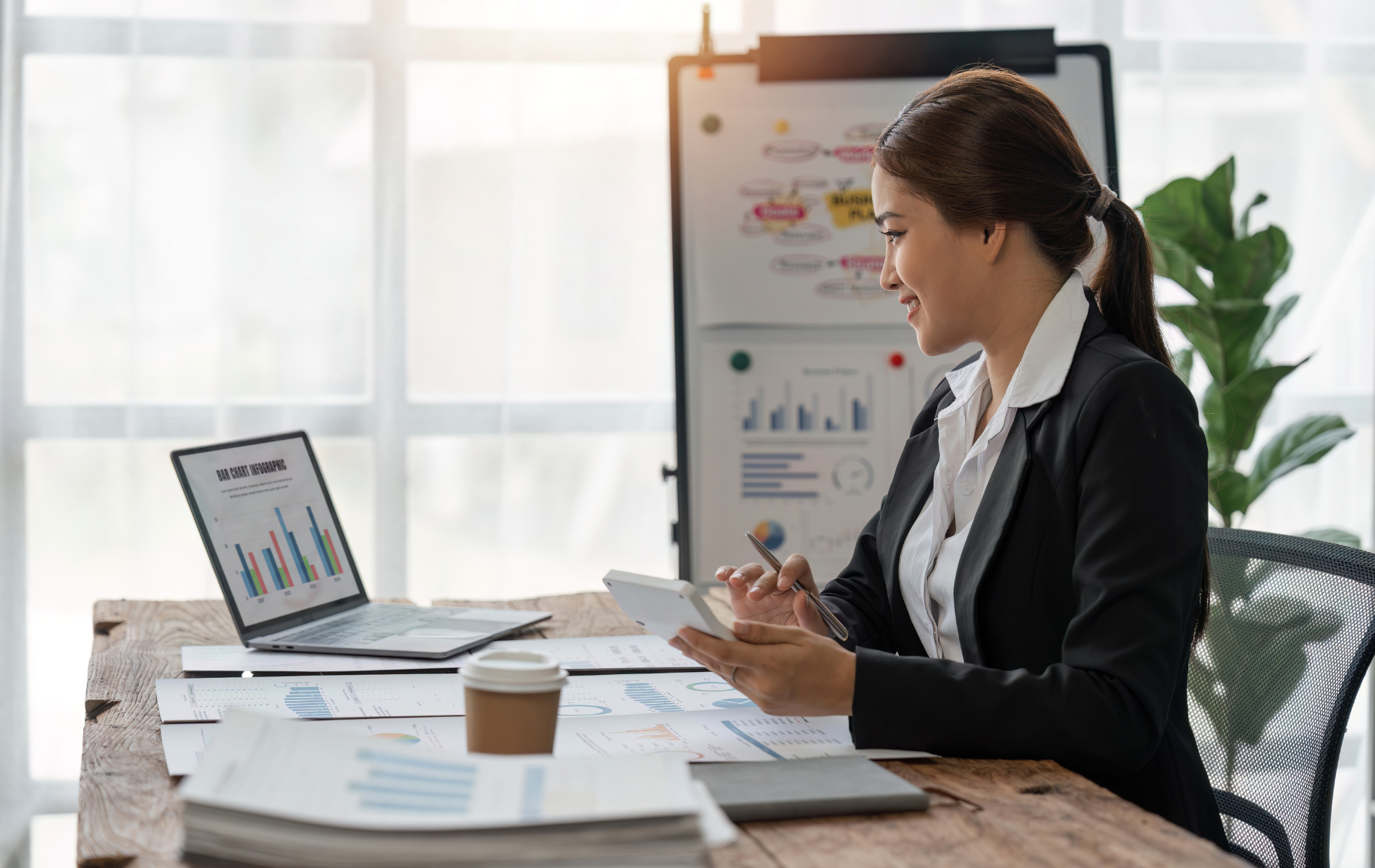Woman in a blazer working at a desk with a laptop, calculator, and charts in a well-lit office.