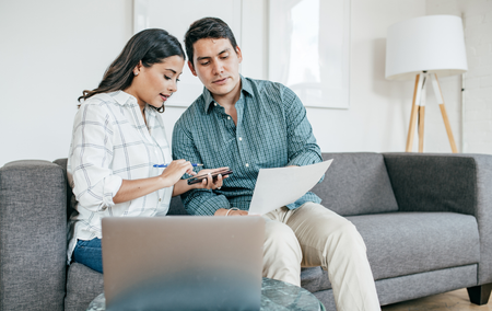 Couple on a couch, calculating finances. Woman points at calculator, man holds papers. Laptop and lamp in background.