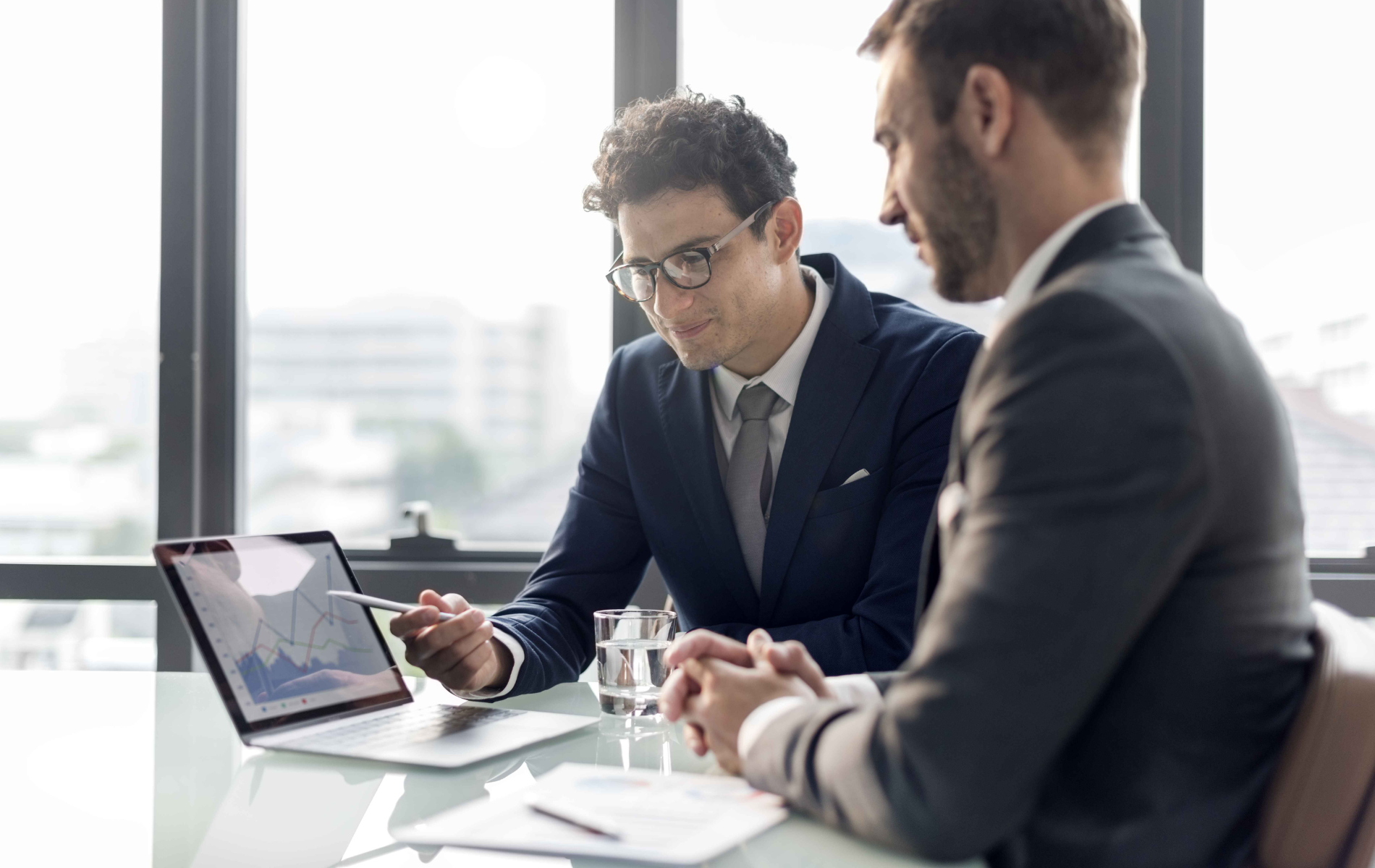 Two businessmen in suits reviewing data on a laptop in a well-lit office.
