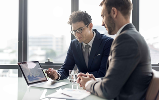 Two businessmen in suits reviewing data on a laptop in a well-lit office.