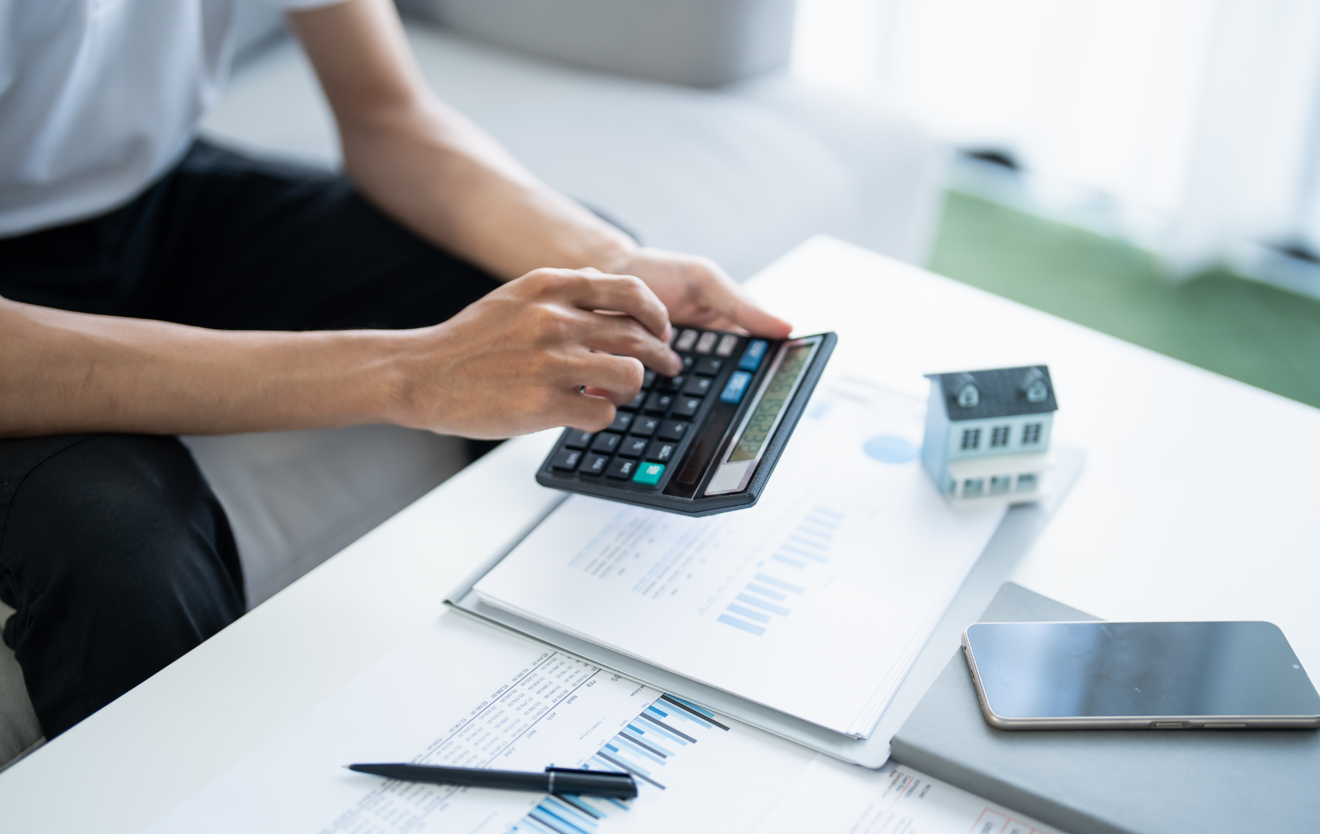 Person calculating on a calculator with a small house model, papers, and phone on a table.