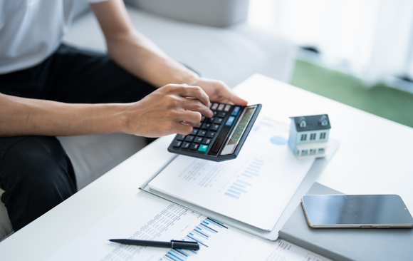 Person calculating on a calculator with a small house model, papers, and phone on a table.
