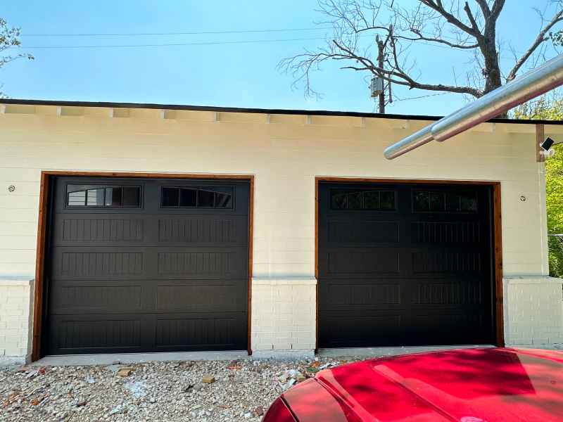 A red car is parked in front of a garage with two black garage doors.