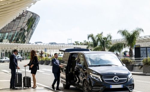 People arriving at a modern building, with a black Mercedes van and luggage carts nearby.