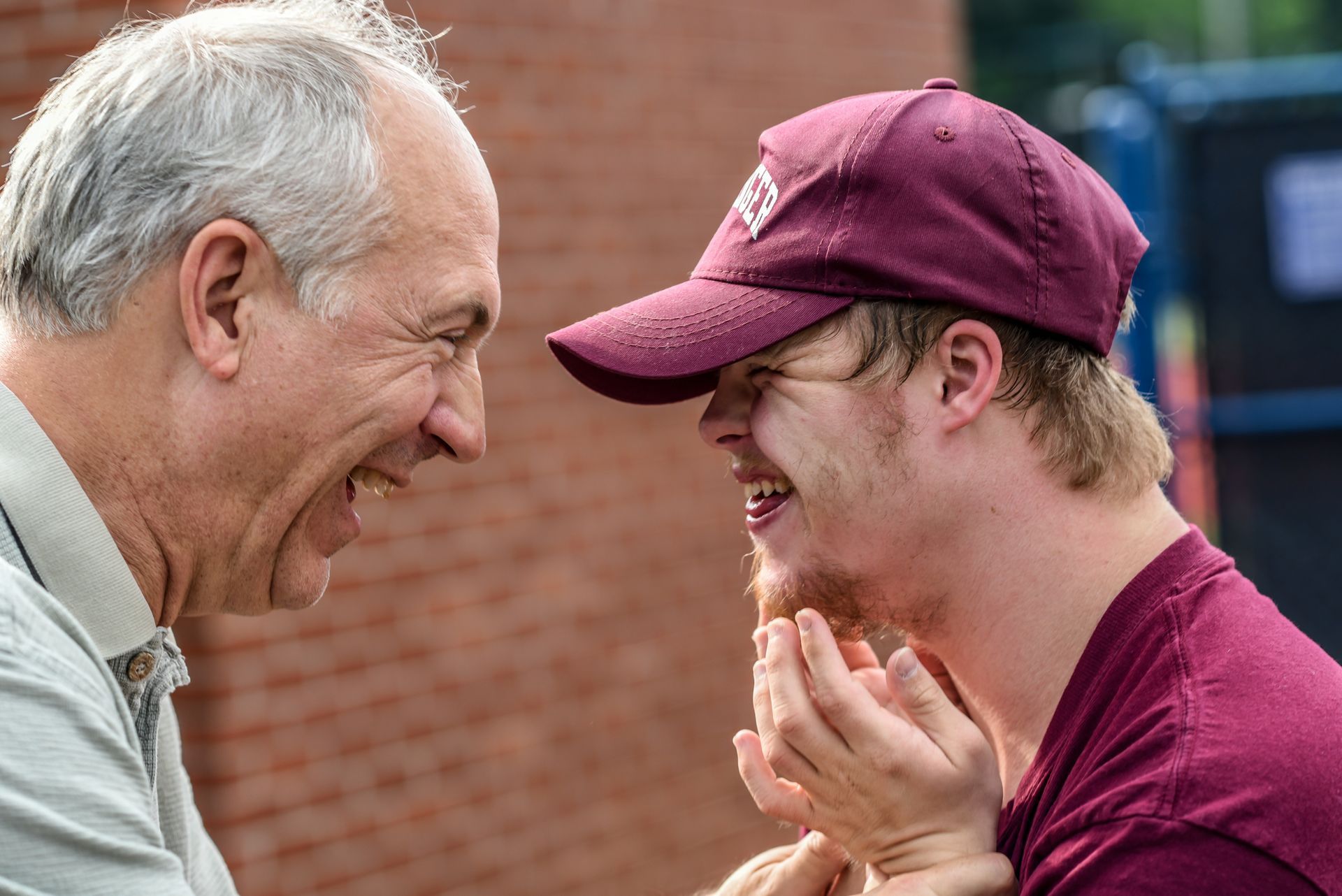 Two people laughing face-to-face outdoors, one in a maroon cap and shirt, the other in a light gray sweater.