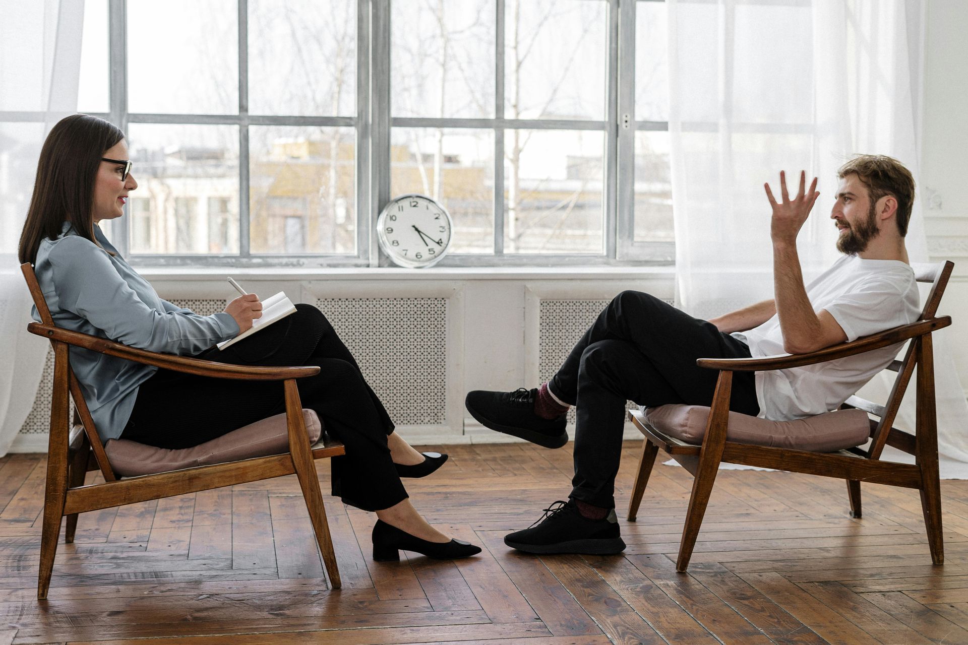 Two people in chairs having a conversation in a bright room with large windows.