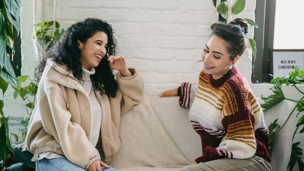 Two women sitting on a couch talking in a bright room with plants and a brick wall