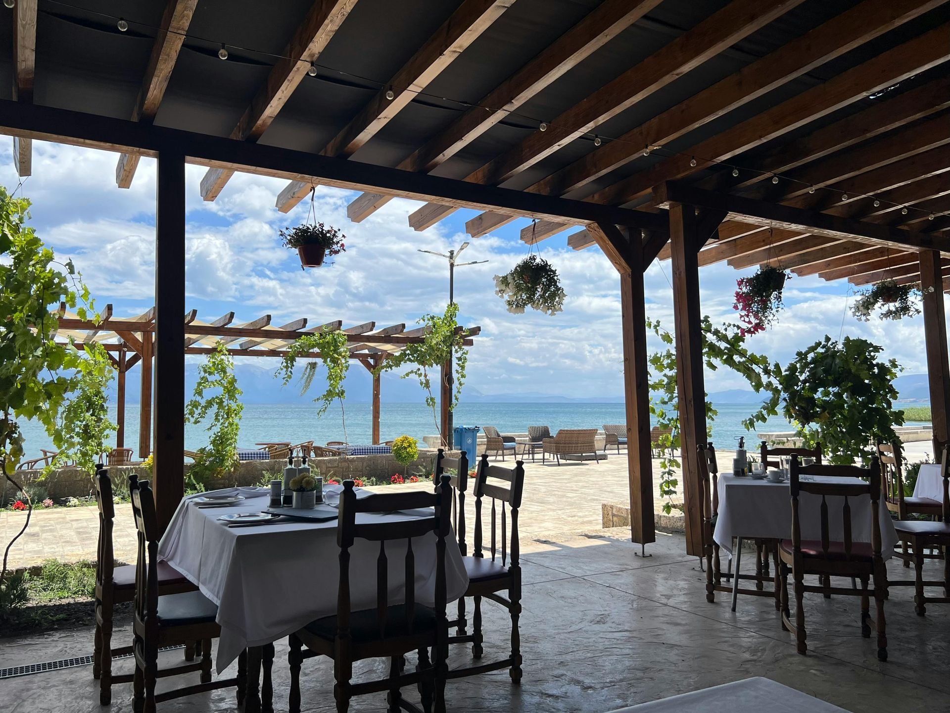 Restaurant patio overlooking a beach with white tablecloths, wooden furniture, and hanging flower baskets.