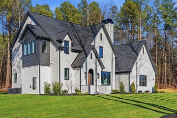 Large white and gray modern house with steep roofs on a green lawn, backed by trees and blue sky