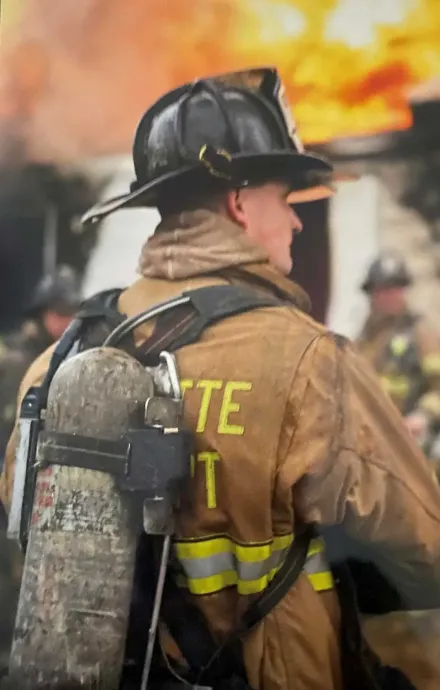 Firefighter in tan gear and black helmet, seen from behind amid flames and smoke.