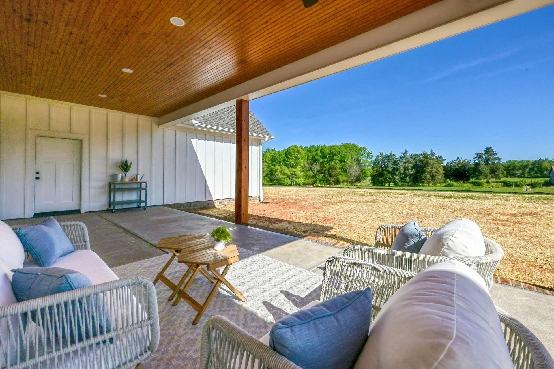 Covered patio with wicker seating and cushions overlooking a sunny field and blue sky