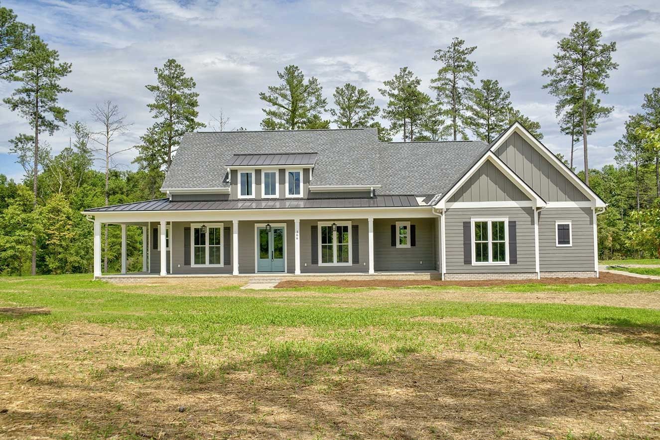 New gray suburban house with front porch and dormers on a grassy lot, surrounded by trees