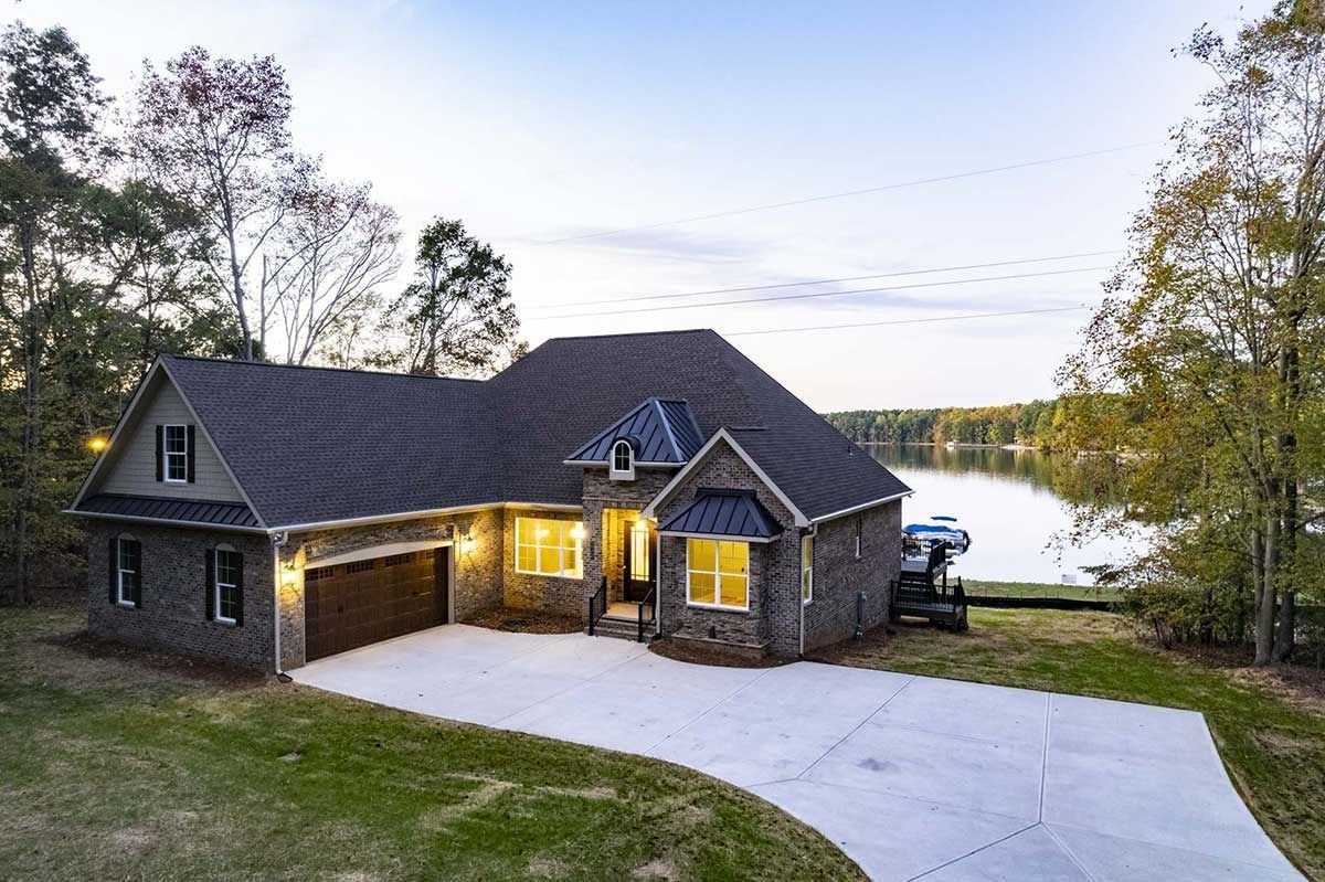 Stone lakeside house with lit porch, gray roof, and white driveway at dusk