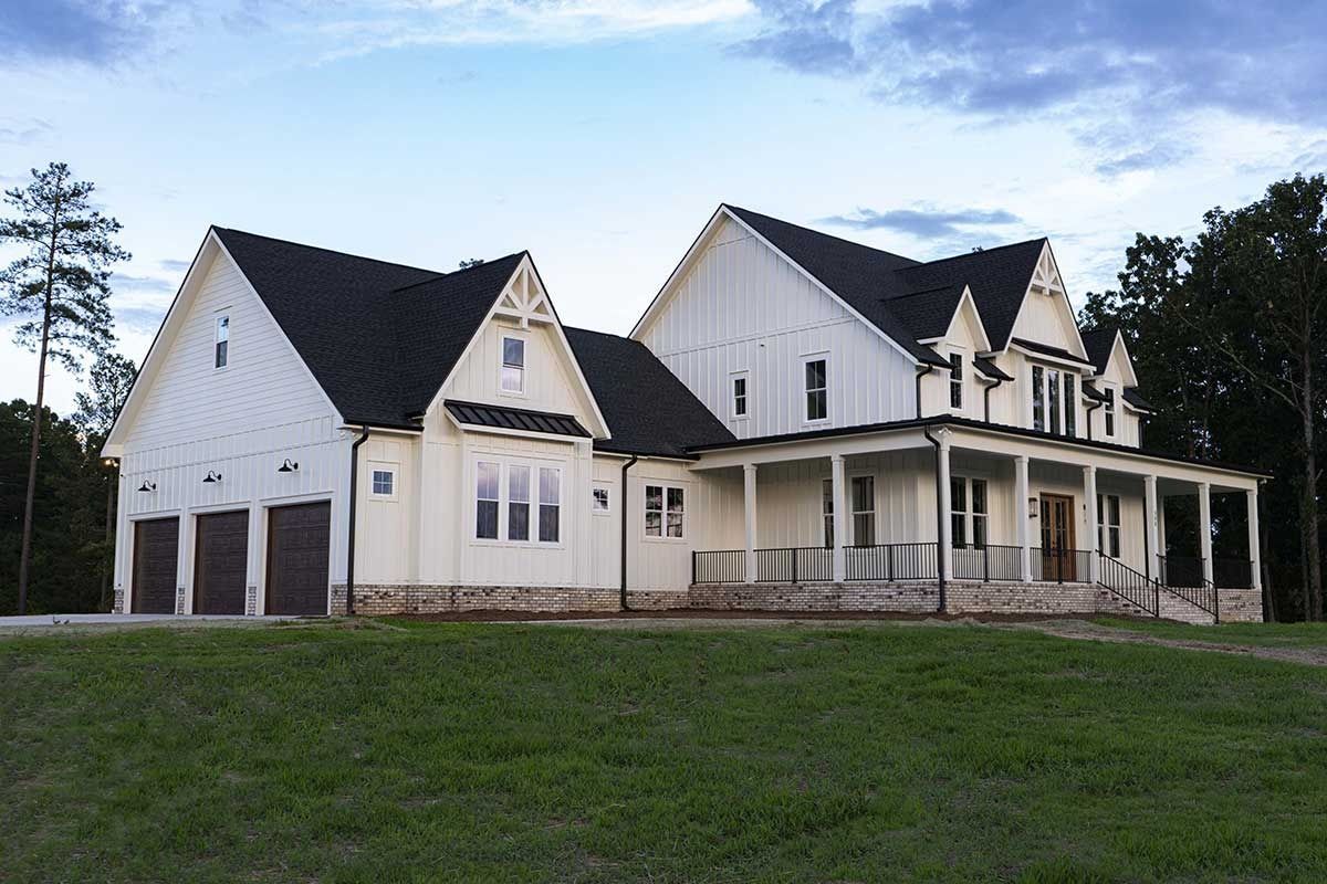 Large white house with dark roof and wraparound porch on a grassy lawn under a cloudy sky