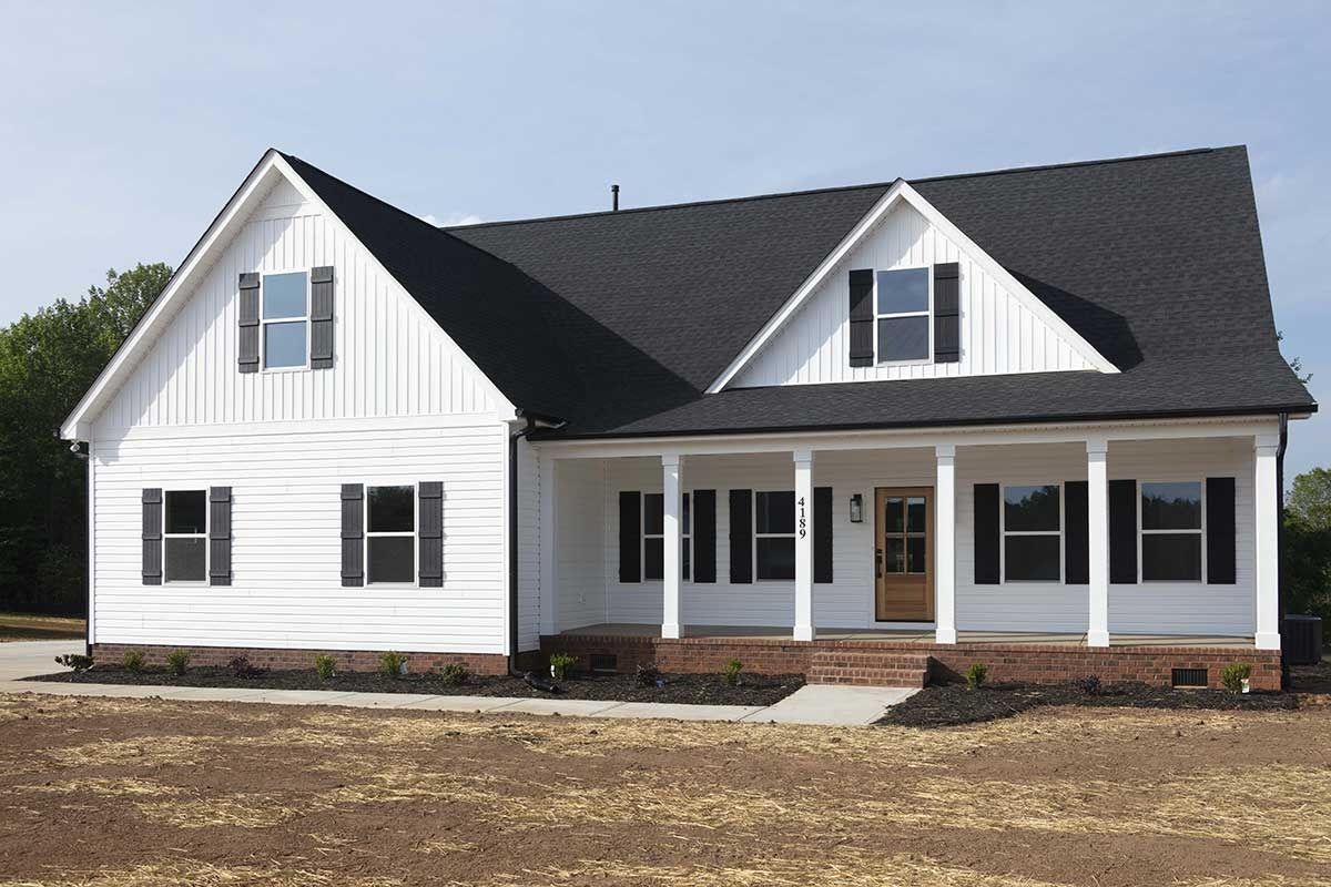 White suburban house with black roof, front porch, and dormer windows on a grassy lot.