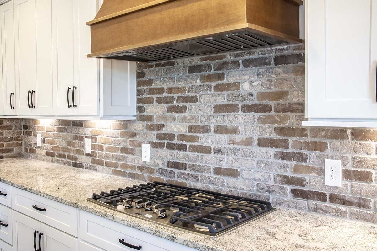 Modern kitchen with white cabinets, brick backsplash, granite counters, and a gas cooktop under a wood hood.
