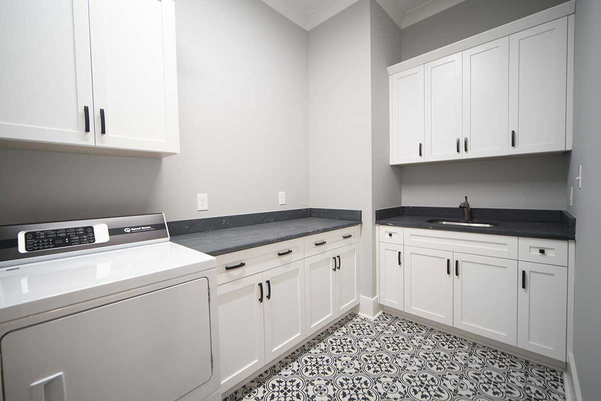 Bright white laundry room with cabinets, dark countertops, washer, and patterned tile floor.