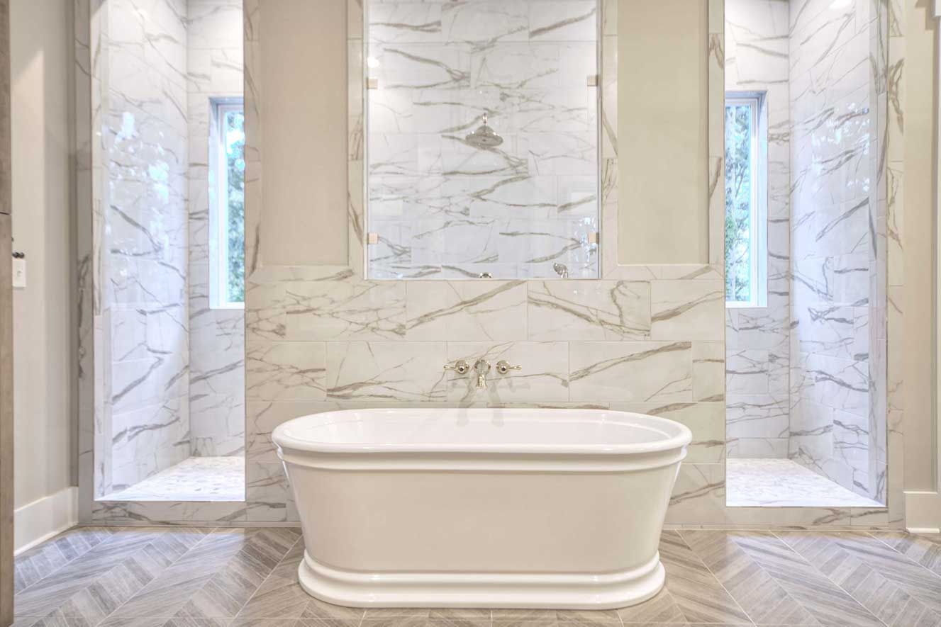 Bright marble bathroom with a freestanding white bathtub centered between two windows.