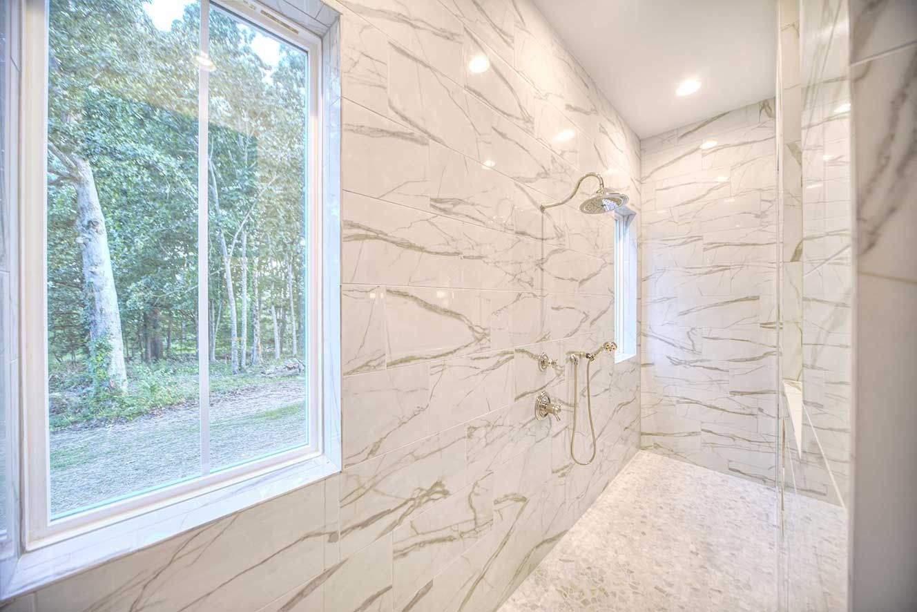 Bright tiled shower with glass window and white marble-style walls, overlooking trees outside