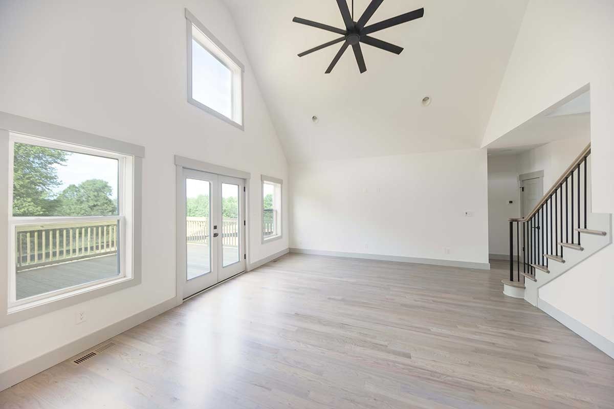 Bright empty living room with high ceiling, black fan, large windows, light wood floors, and staircase